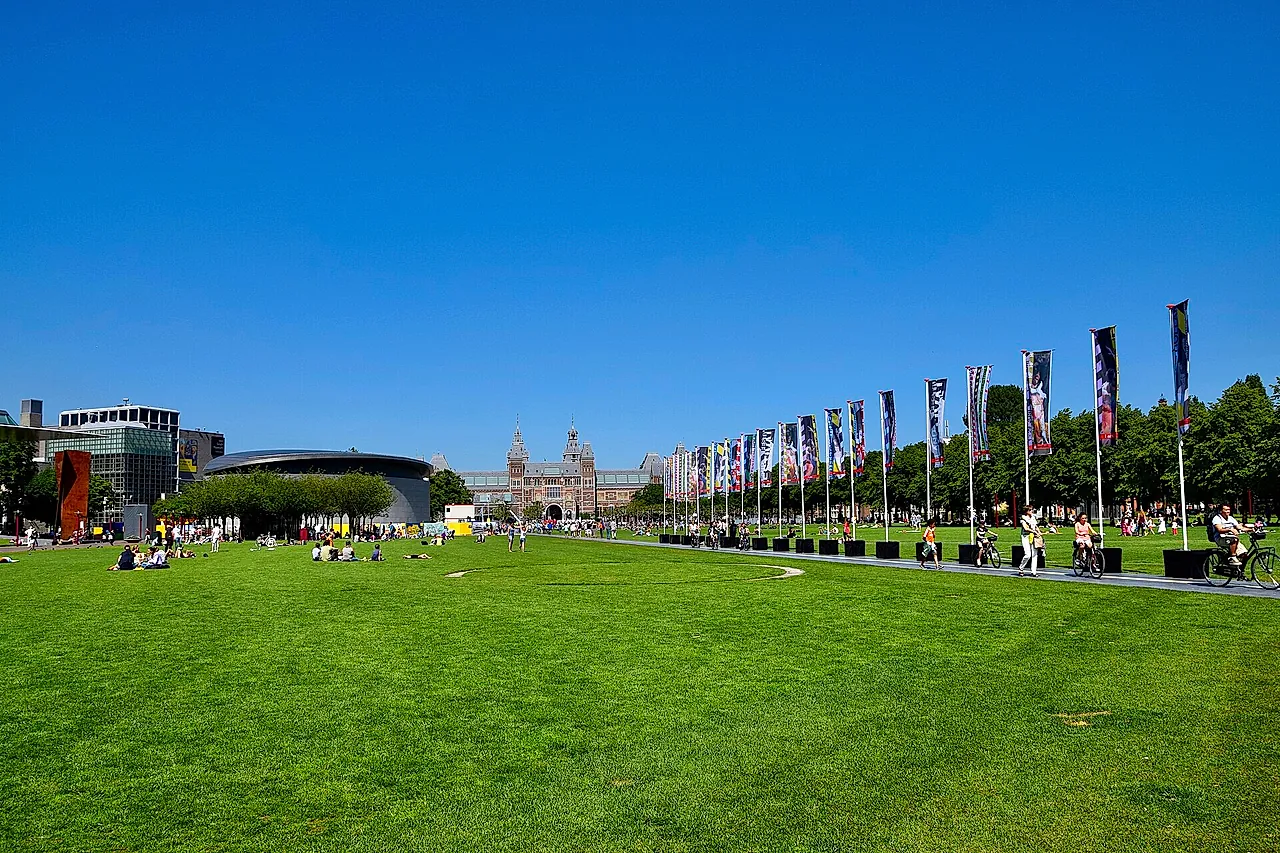 The wide lawn of Museumplein under a blue sky, with a row of flags and the Rijksmuseum and Van Gogh Museum on the horizon